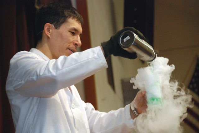 jeff evans magician, in a white shirt pouring something from a dark bottle into a foaming beaker
