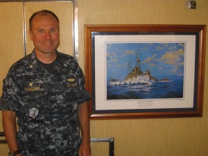 Photo of Commander Tom Shugart, Commanding Officer of the USS OLYMPIA standing next to a painting of the Cruiser Olympia. 