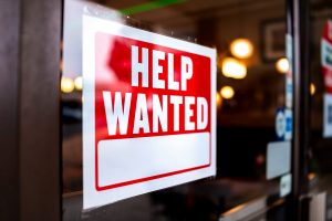 Sign text closeup for help wanted with red and white colors by entrance to store shop business building during corona virus covid 19 pandemic