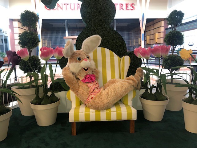 A person dressed as the Easter Bunny laying on a yellow striped chair with a bunny topiary behind them.