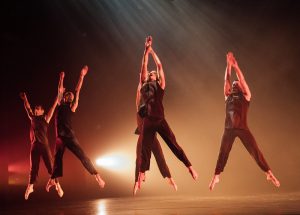 Group of five dancers on a black stage with bright lighting