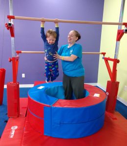 a teacher at The Little Gym helps a child reach for mini uneven bars