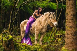 woman in mermaid costume on a fjord horse with a unicorn horn in a forest