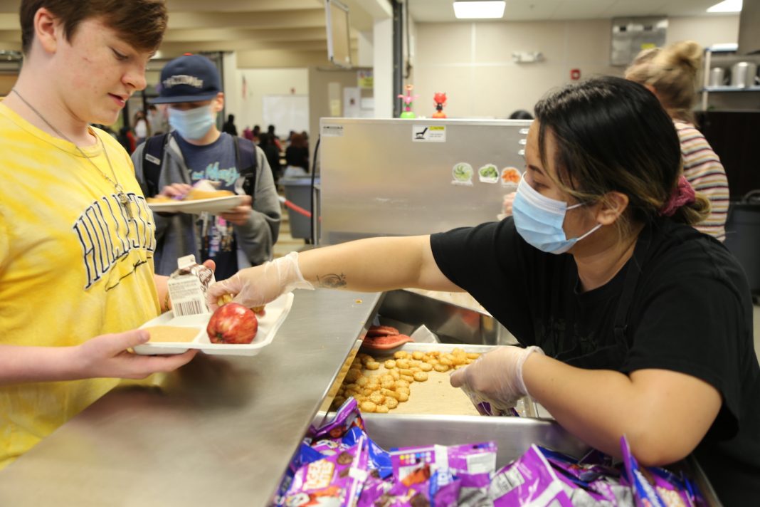 child in mask at a school cafeteria being handed food by a lady in a mask