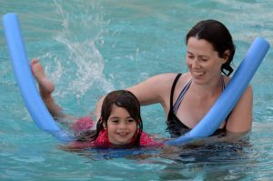 girl on a pool noodle in a pool with an adult