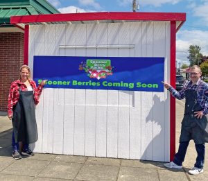 white with red trim wooden farm stand with Spooner Farms name and two people standing on either side