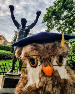 Western Governors University's mascot Sage, a stuffed owl, in front of a statue