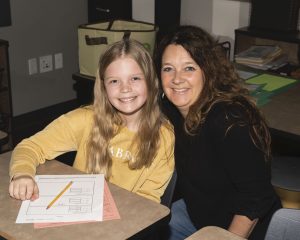 a student at a desk with a teacher kneeling next to her, both smiling at camera