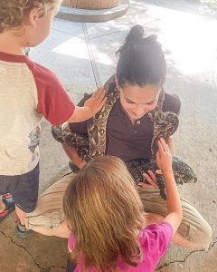 lady kneeling down with a large snake around her neck, two children are petting the snake