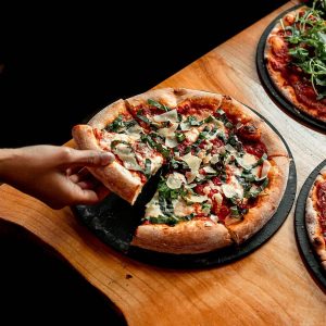 person's hand taking a slice of pizza from a table at Well 80