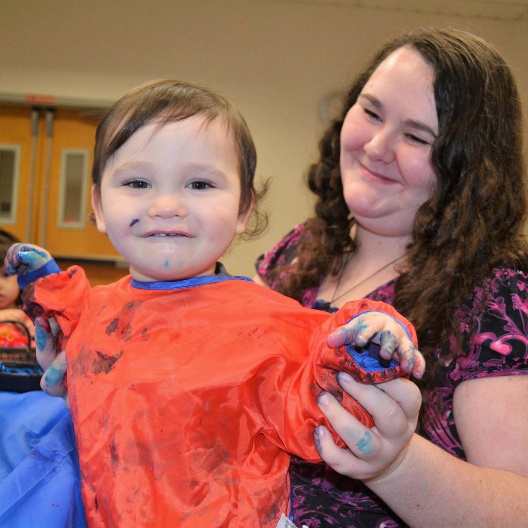 young child smiling while a woman holds her hands, covered in paint, up