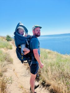 Kris Dales at the ocean with a baby on his back.