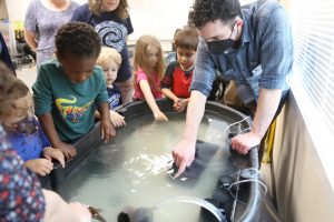 un groupe d'enfants avec un enseignant regardant à l'intérieur d'un grand bac en caoutchouc noir rempli d'eau et de filtres