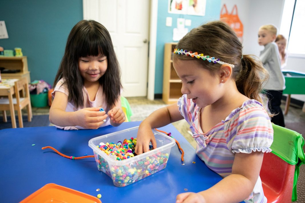 two preschool girls sitting at a table sifting through a container of beads