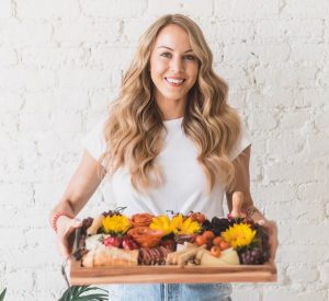 woman holding a wooden tray full of flowers, cheese, bread, salamis and other meats