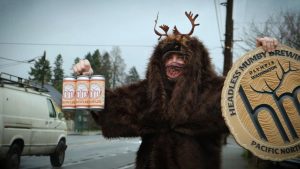 Person in a furry coat with antlers holding a six pack of beer and a Headless Mumby Brewing sign