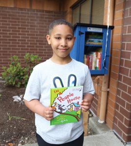 young student holding a book next to a Little Free Library