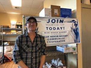 Evan Price, owner of the Blue Heron Bakery standing in front of a banner announcing the co-op at Blue Heron Bakery