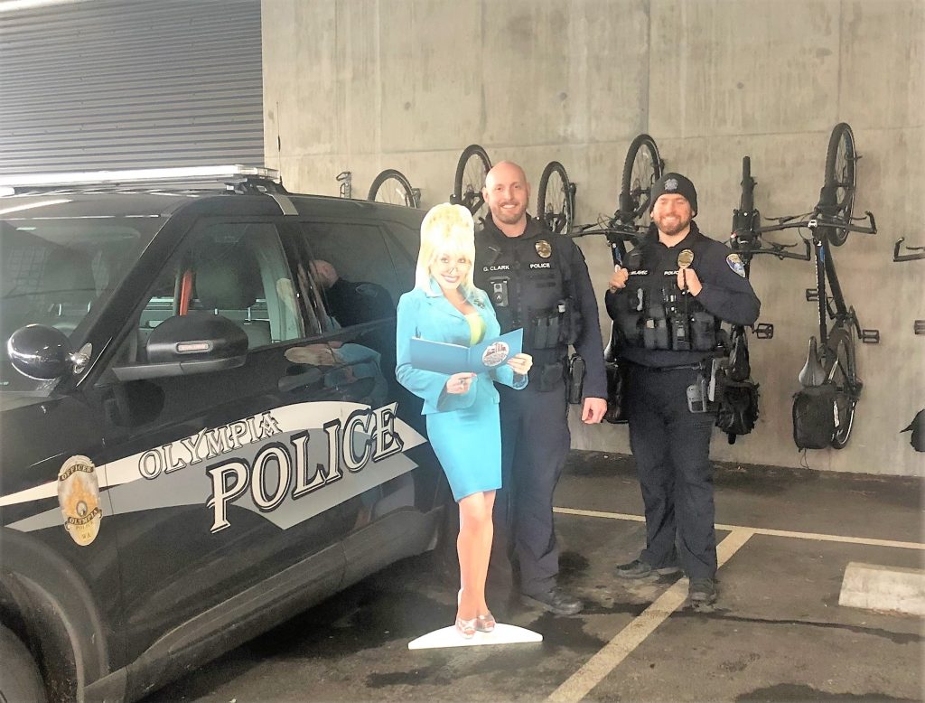 cardboard cut-out of Dolly Parton standing by a Olympia police care and a few officers