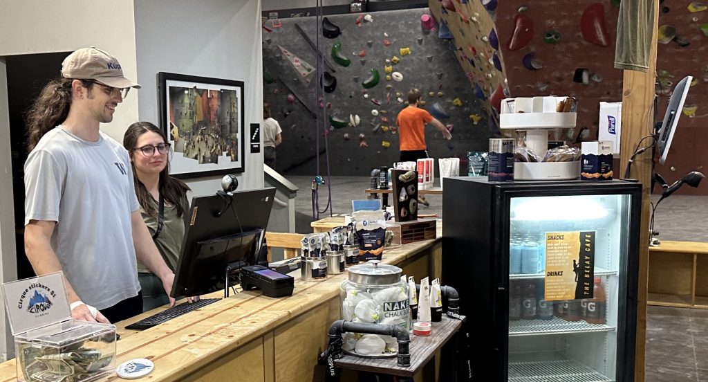 two people standing at the front desk at the Cirque Climbing Gym, with a glass door-fridge in front with energy drinks and other products on the counter