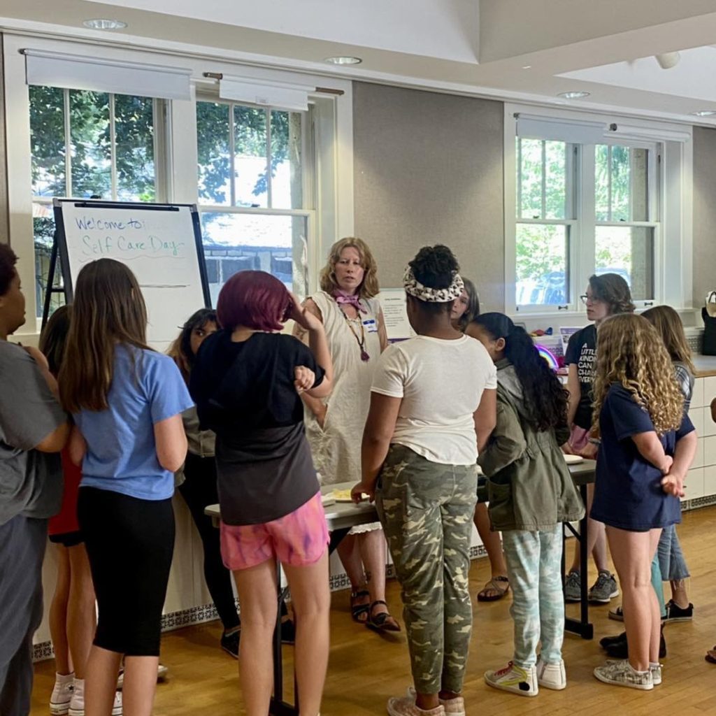 Group of girls standing at a table and listening to a woman talk