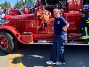 Maggie Bean in jeans a fire house shirt standing by a vintage fire truck