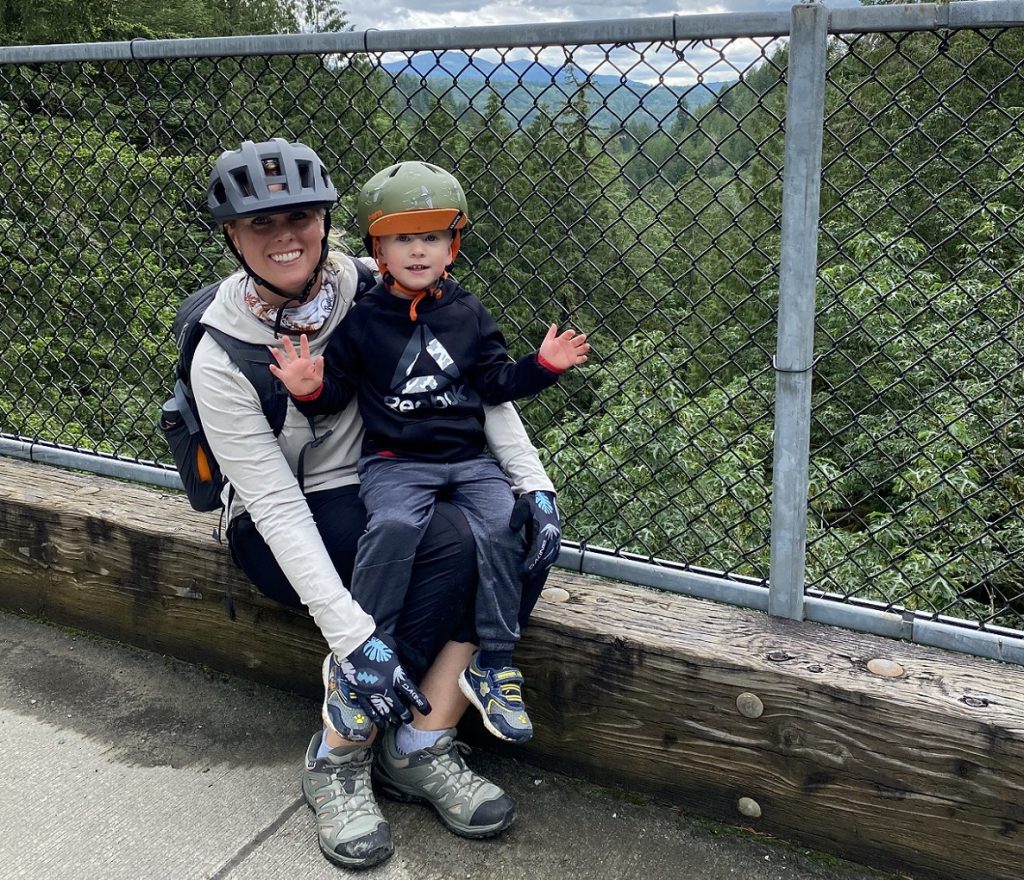 Christine Hammer kneeling with a kid on her lap against a chain-link fence overlooking a forest, both wearing bicycle helmets