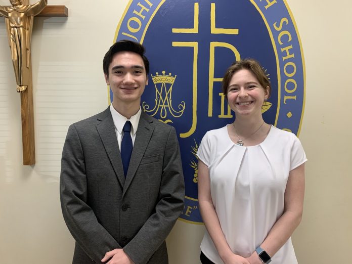 Nathan Miller and Catelin King standing in front of the Pope John Paul HIgh School sign with a crucifix on the wall to the left of Nathan.