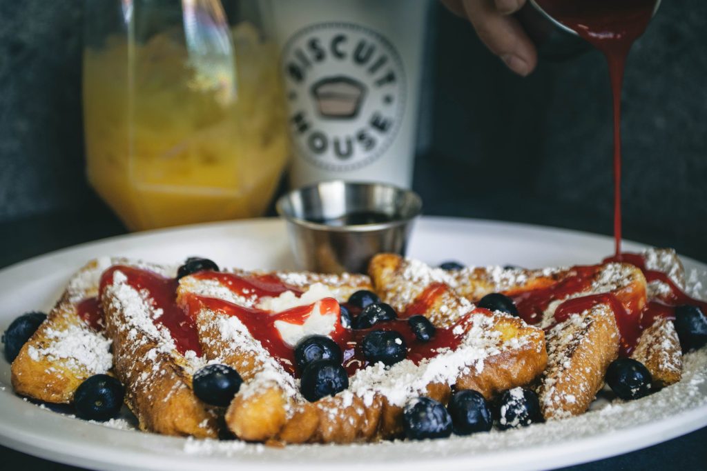 French toast with blueberry, powdered sugar, butter and red berry syrup being poured over it.