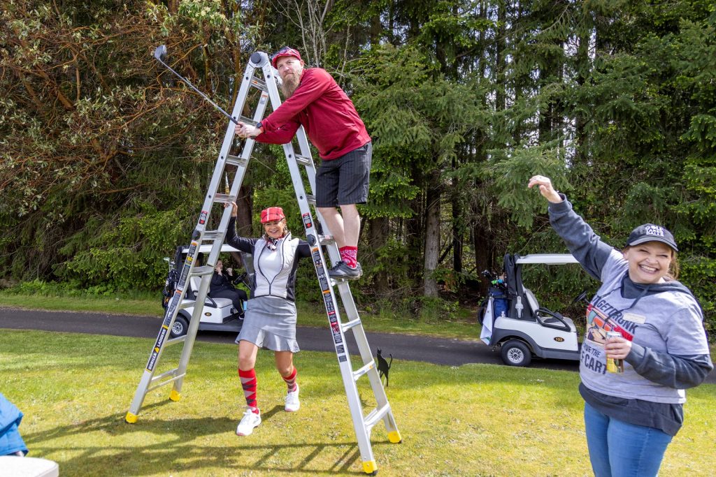 Golfer standing on a metal ladder hitting a golf ball with another golfer standing underneath it and someone else standing next to it cheering