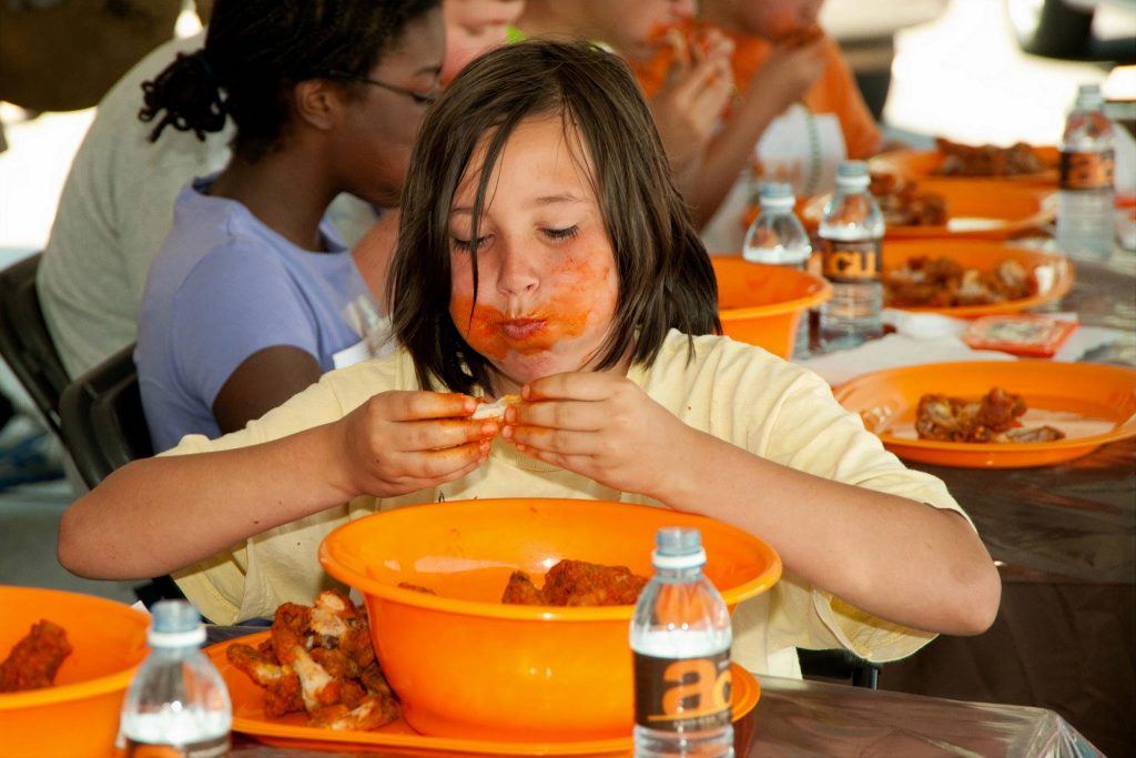 kid eating a bunch of chicken wings out of an orange bowl, face is covered in bbq sauce