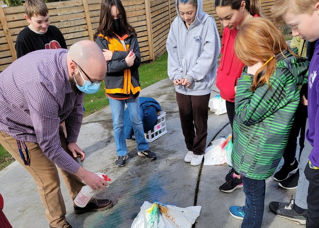 Students standing around a teacher who is spraying colored liquid on a white piece of paper outside