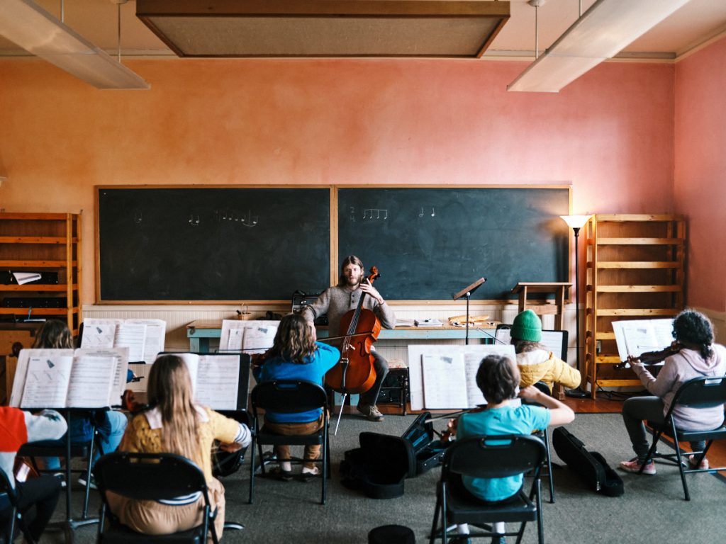 A music teacher playing the cello with students on their string instruments with music in front of him