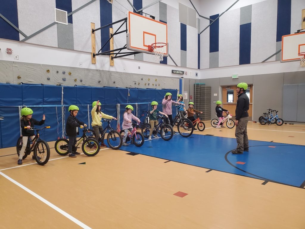 kids in a gym with bikes and helmets and an instructor standing in front of them
