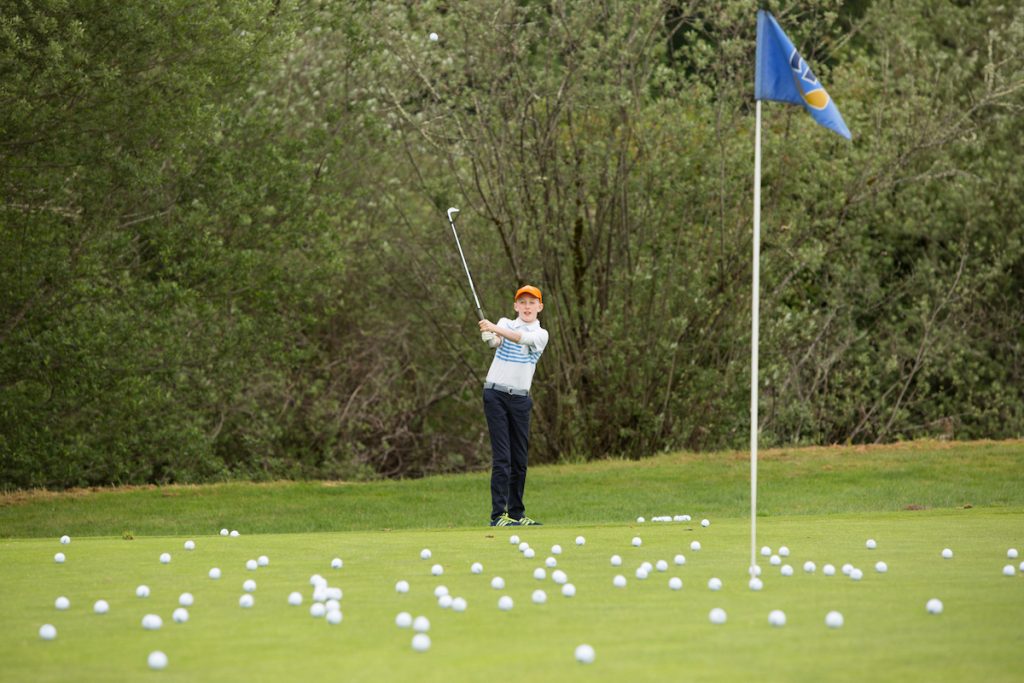 a youth swings a gulf club in the background with white golf balls scattered on the green by the hole.