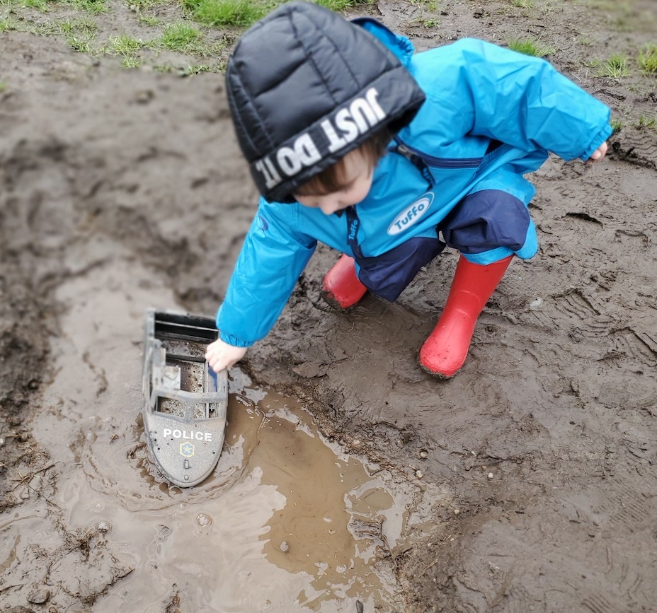 child in rain gear playing with a toy boat in a mud muddle