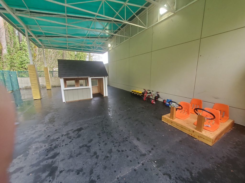 Covered outdoor space with rubber mats on the ground, a play house, trikes and other toys