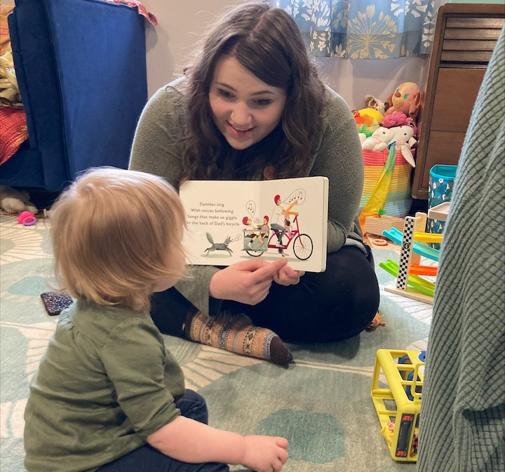 a woman sitting on the floor with a toddler. The woman is holding a book open, faced toward the toddler