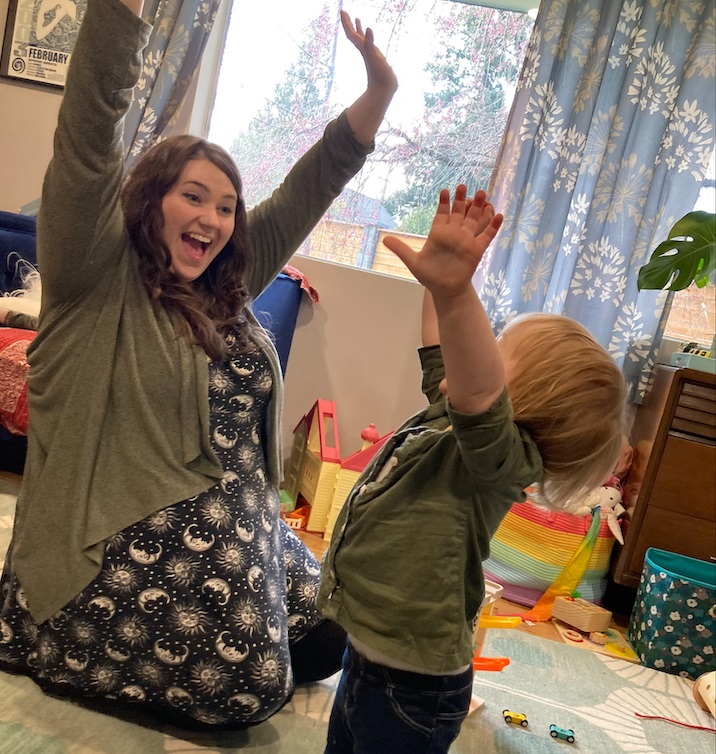 CHERISH Co-lead Marisha Bruce sitting on the floor with her hands up in the air smiling with a toddler standing with his hands up in the air smiling