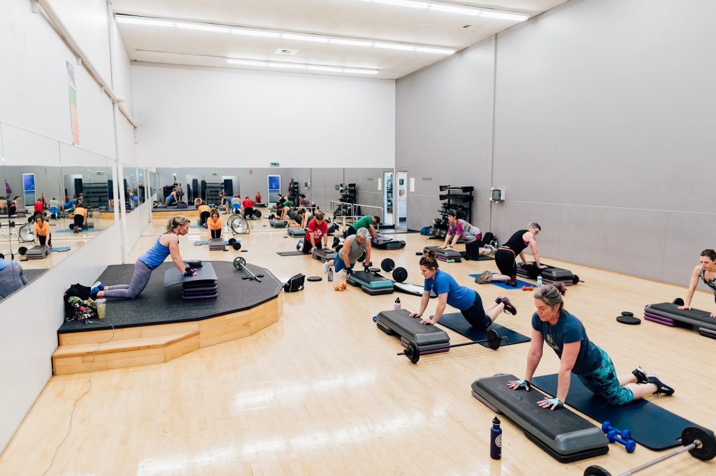 group of people in a gym room with mirror with an instructor kneeling with hands up on a large stepping mat and people in the class doing the same