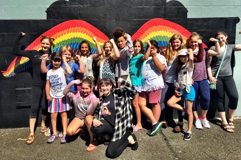 large group of young women standing in front of a rainbow mural