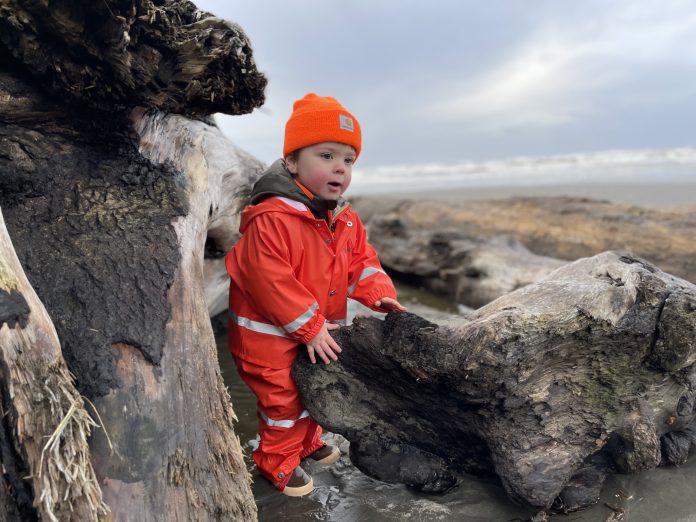 Washington Coastal Marine Sanctuary WA Coast little boy in a orange rain suit and orange beanie stands among driftwood on an Olympic beach.