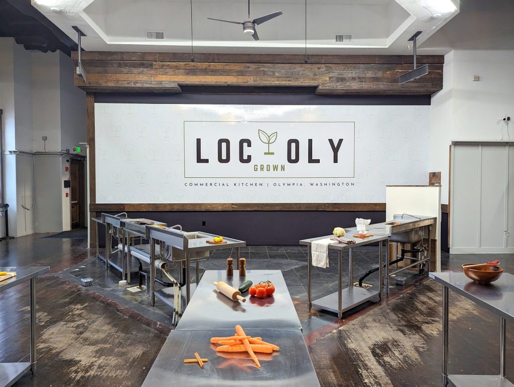 metal industrial kitchen counters with carrots, rolling pin, tomatoes, a cucumber and other items on them. Wall says 'Loc-Oly Grown. Commercial Kitchen, Olympia, Washington'