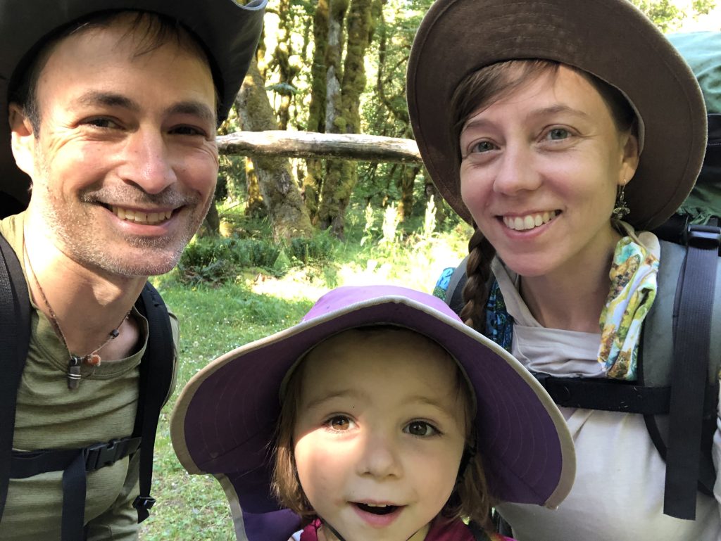 Chris and Tiffany selfie with a little girl in a hat in-between them standing in a woods.