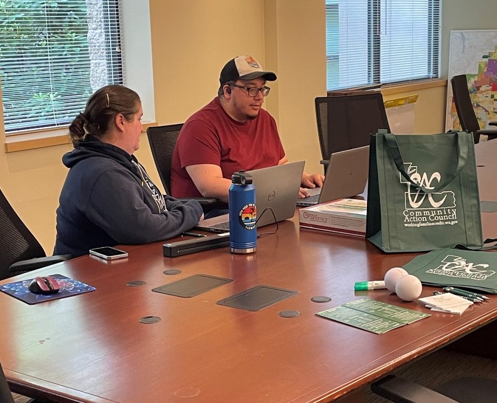 two people sitting at a large table with swag bags and a laptop on it