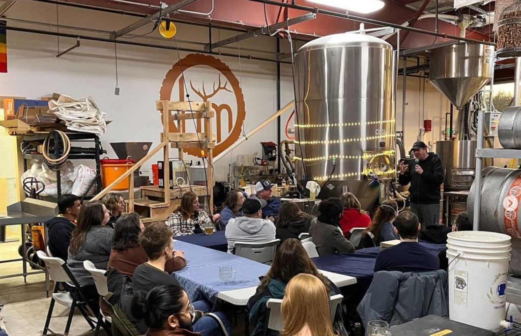 Large group of people sitting at folding tables inside Headless Mumby Brewing with a man speaking into a microphone