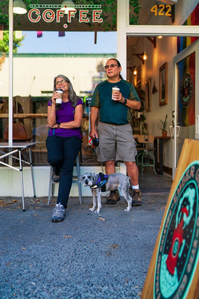 two people and a dog standing outside Ember Goods with to-go coffees