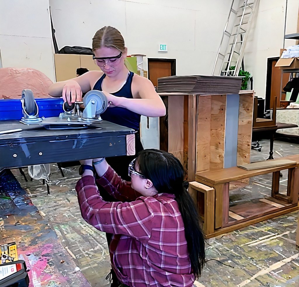 Two women, one standing, one kneeling, attaching a set of wheels to a black wooden platform