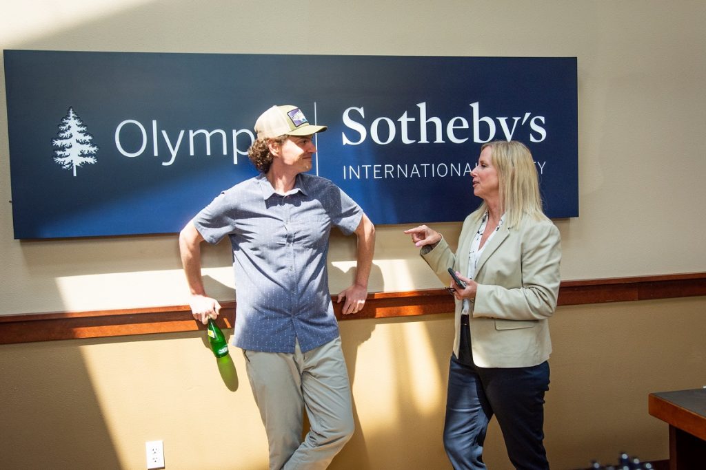 a man and a woman staand talking against a wall with a blue 'Olympic Sotheby's International Realty' sign.