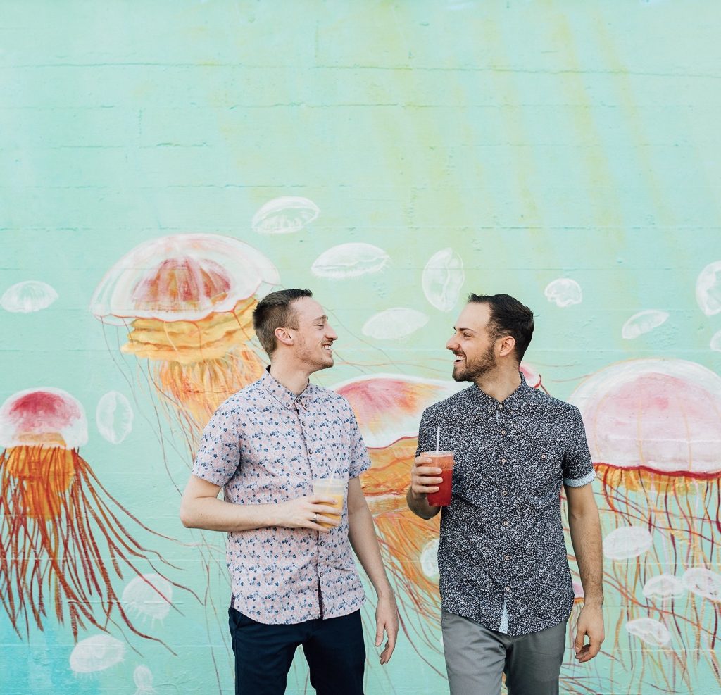 Konrad J. Bruns-Rytting and Nate Thomas  smiling at each other while holding drinks in front of a jellyfish mural